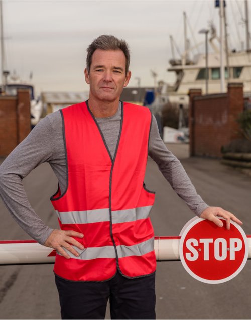 Gilet R200 rouge personnalisable dès 1 pièce Homme portant un gilet R200 rouge à 2 bandes réfléchissantes devant une barrière de contrôle d'accès